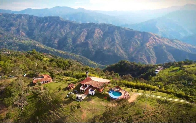An aerial view captures a charming property nestled in lush greenery, surrounded by rolling mountains. A swimming pool is visible, alongside multiple open spaces. The structure features a traditional roof, blending harmoniously with the natural landscape.