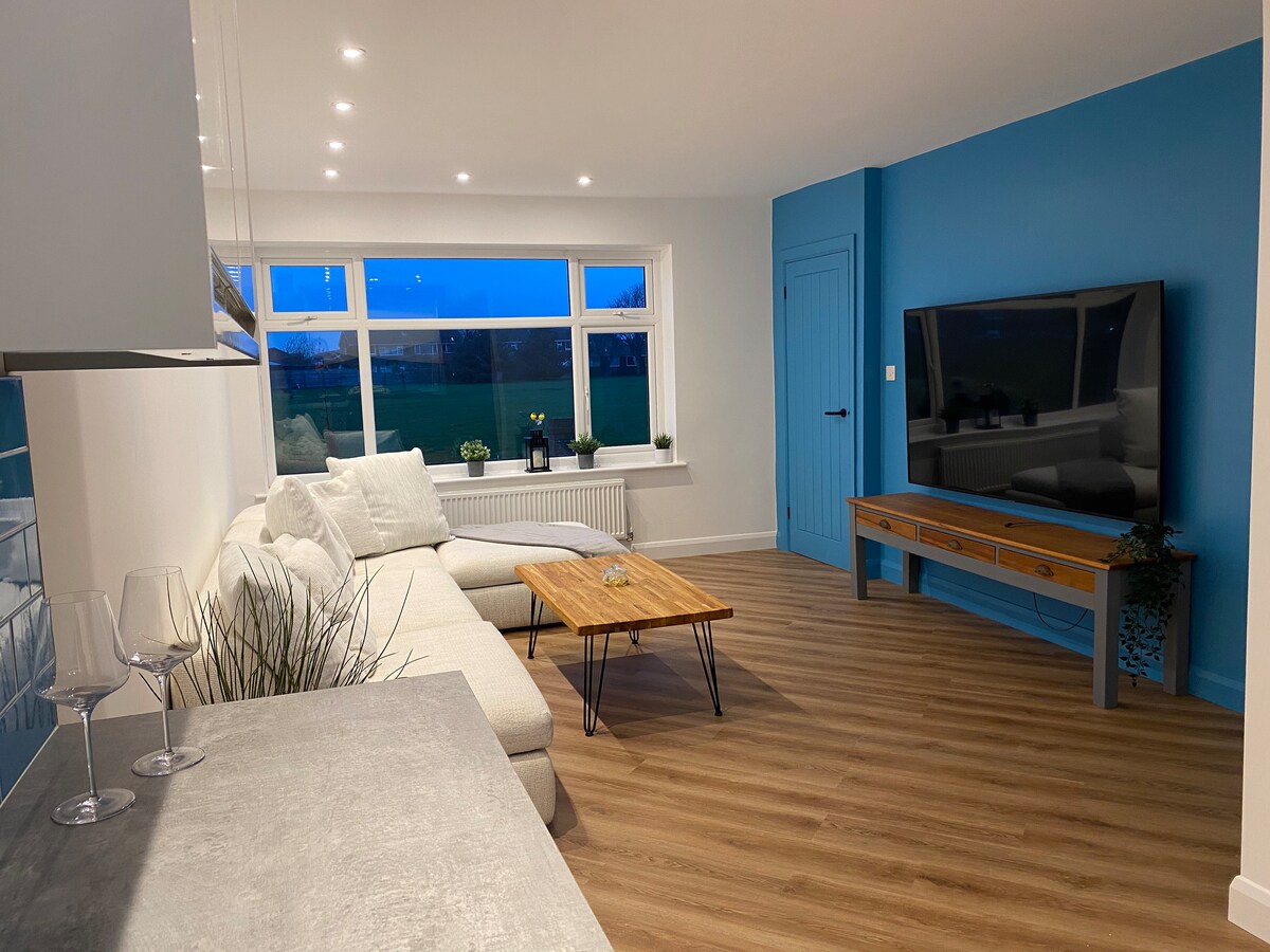 A seating area features a light-colored sectional sofa and a wooden coffee table, set against a vibrant blue accent wall. Natural light enters through the large windows, and decorative plants complete the serene environment. A flat-screen television is positioned on a wooden console.