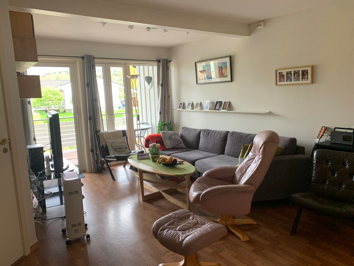A light-filled living area features a large gray sofa and a small round coffee table topped with fruit. In the corner, a comfortable armchair sits beside a television stand. Large glass doors open to a balcony, allowing views of greenery outside.