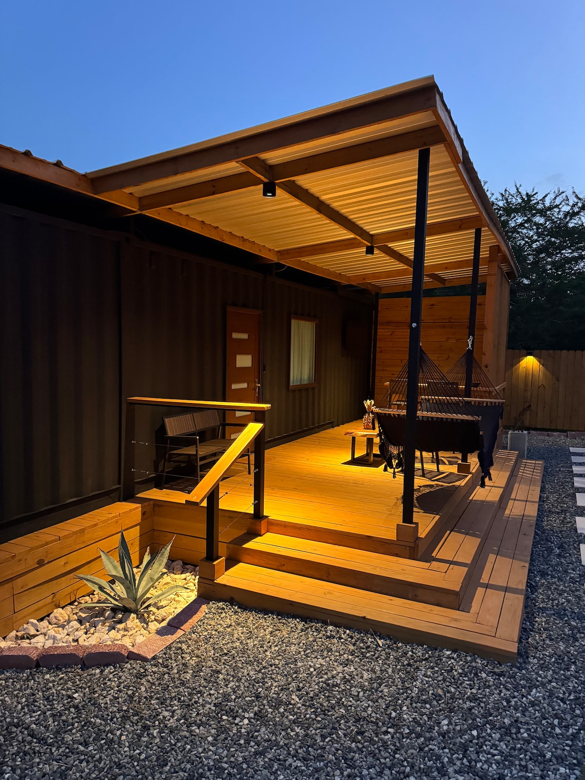 An outdoor terrace deck is illuminated by warm lighting, featuring wooden steps leading up to a cozy seating area with two chairs. A nearby plant adds a touch of greenery to the gravel walkway, while the container home's exterior walls provide a rustic backdrop.