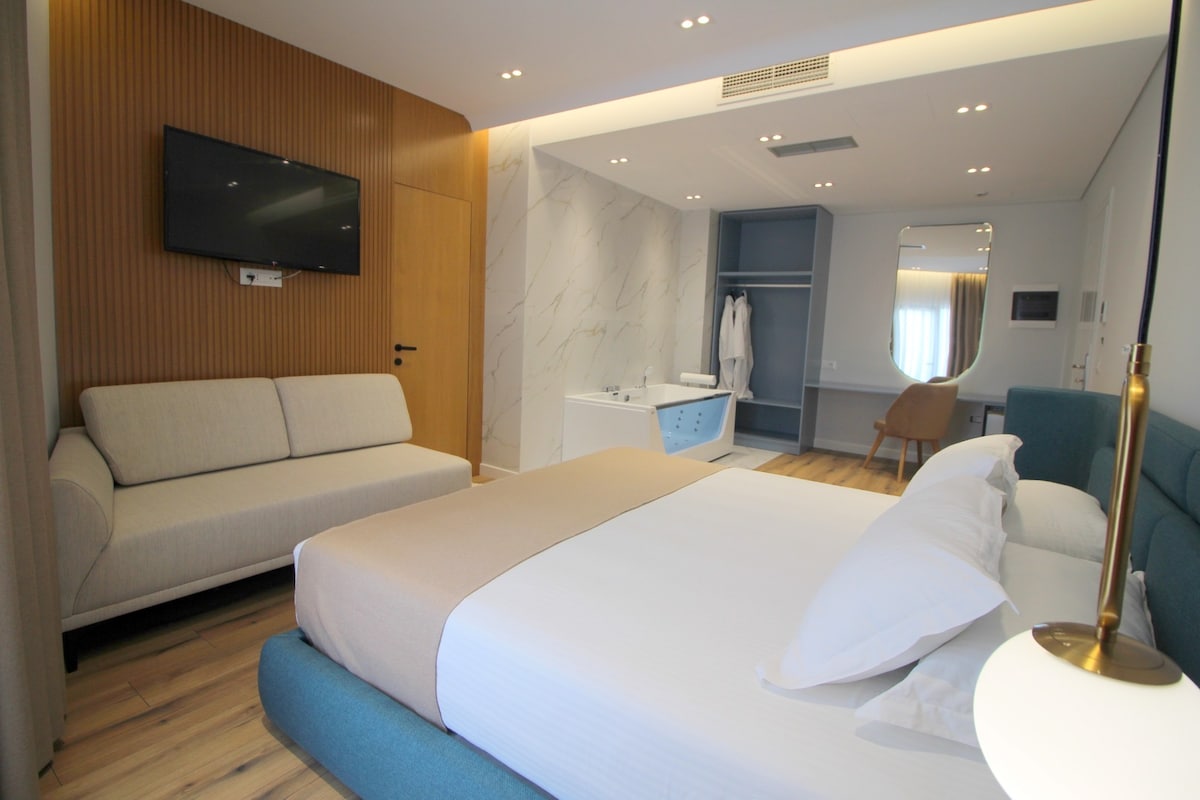 A contemporary room is shown, featuring a spacious bed with neutral-toned bedding. A sleek, light-colored sofa is positioned nearby, while a modern built-in wardrobe and wash area are visible in the background. Soft lighting complements the wood and marble accents throughout the space.