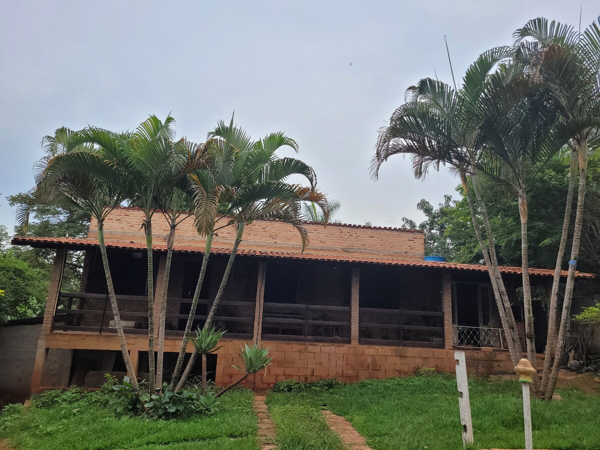A rustic building with a red-tiled roof is surrounded by lush greenery. Tall palm trees stand in front, and a pathway leads up to the entrance. Large open spaces with wooden railings enhance the inviting appearance of this group-friendly accommodation.