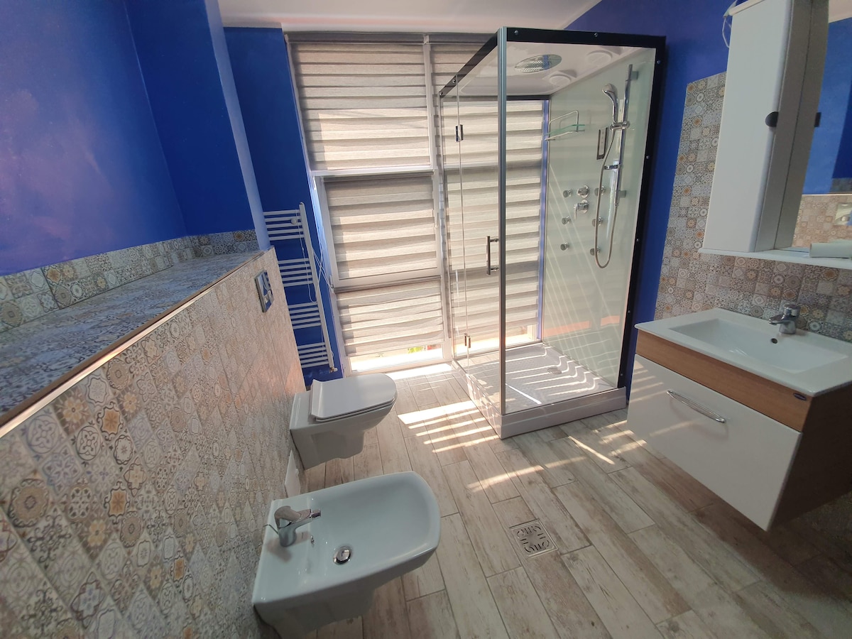 A modern bathroom features a walk-in shower enclosed by glass. A wall-mounted sink and storage cabinet are present. Two toilets are positioned beside decorative tiled walls. Natural light enters through the large window, with blinds partially drawn, creating a bright space.