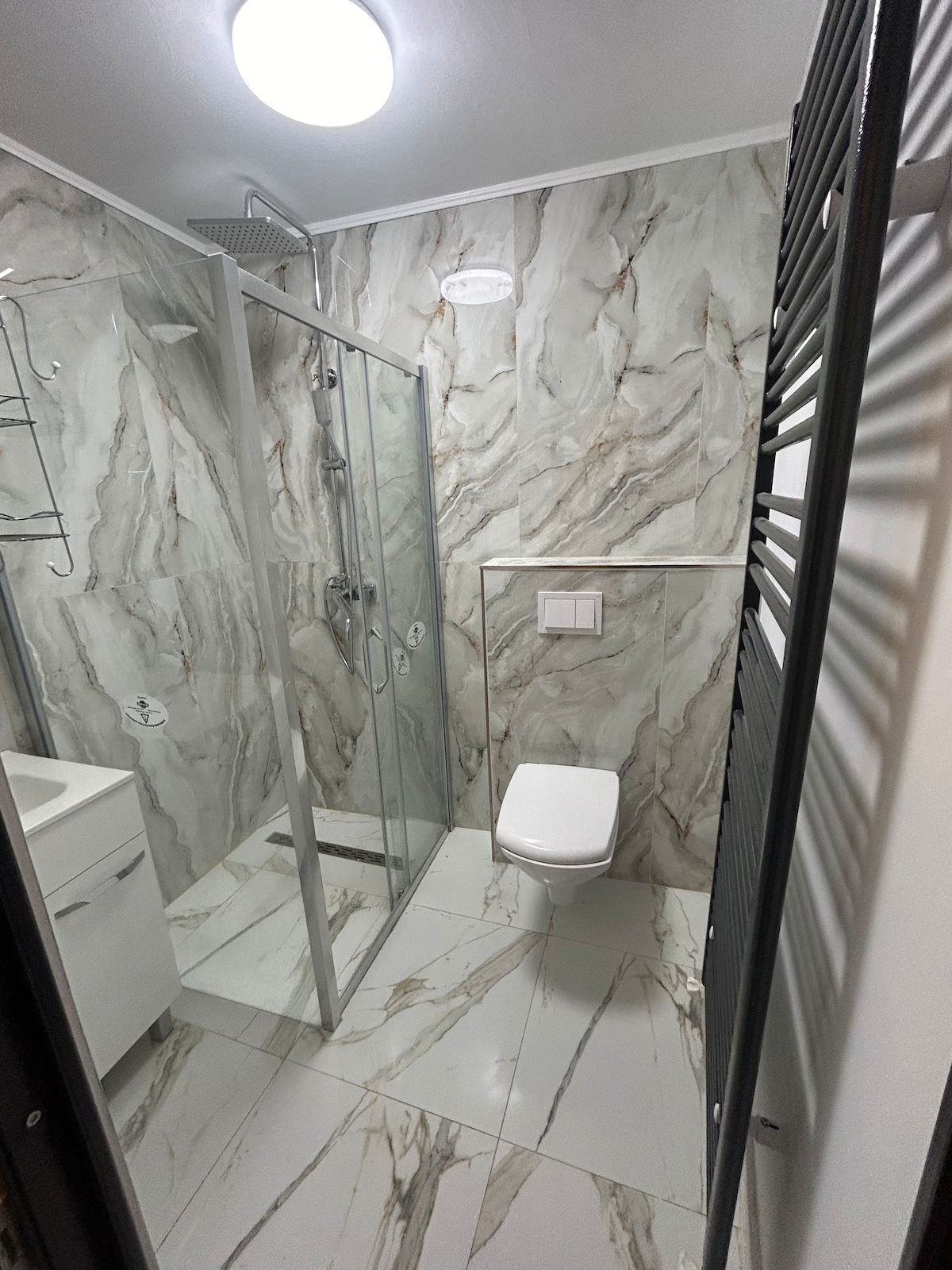 A modern bathroom featuring elegant marble walls and floor tiles. A glass shower enclosure is installed alongside a sleek toilet. A minimalist vanity with a storage unit is present, complemented by neutral tones and soft lighting.