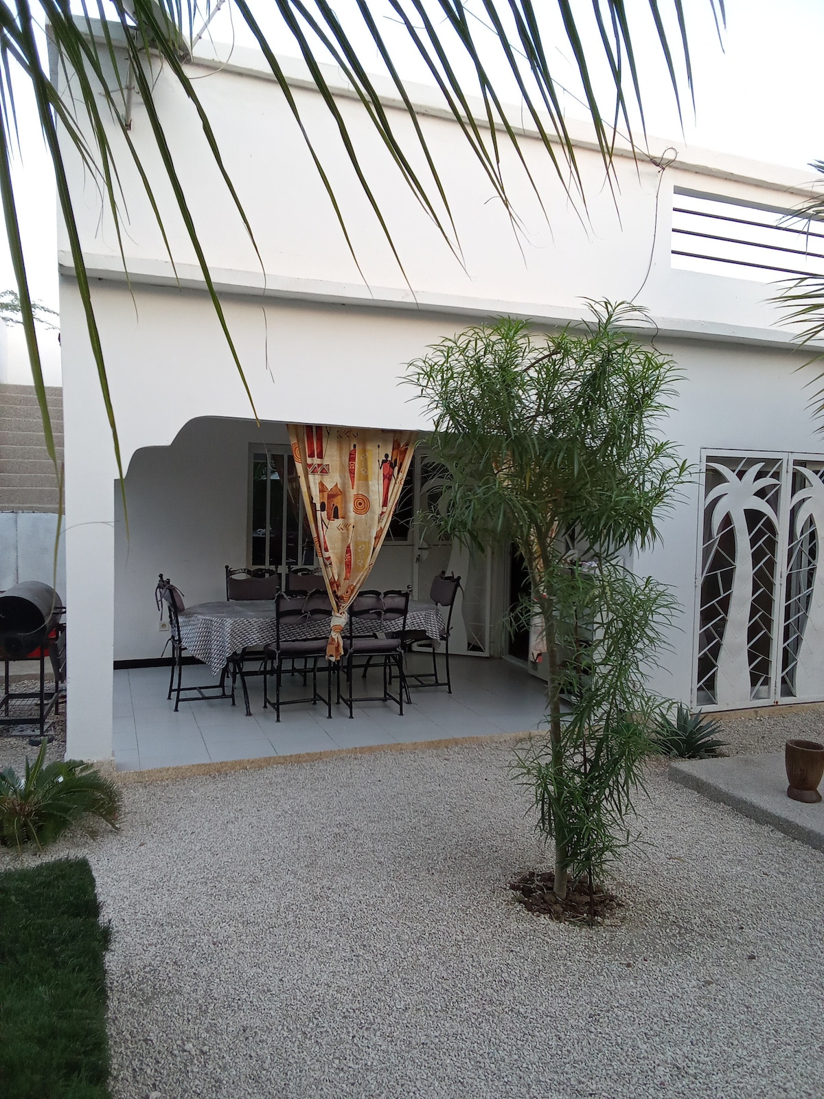 An outdoor dining area is featured under a covered terrace, with a large table set for dining. Surrounding greenery includes a small palm tree and well-maintained garden gravel. Light-filtering curtains in a colorful design enhance the relaxed atmosphere of the space.