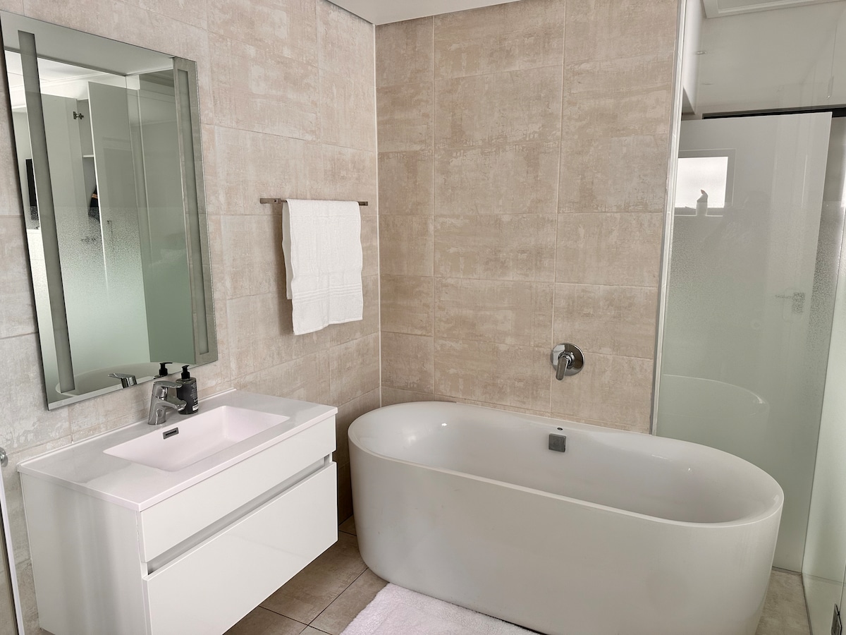 A modern bathroom features a freestanding oval bathtub, complemented by a sleek vanity with a minimalist design. A large mirror reflects light, enhancing the space. A glass shower enclosure is visible, and plush white towels are neatly hung on a rack.