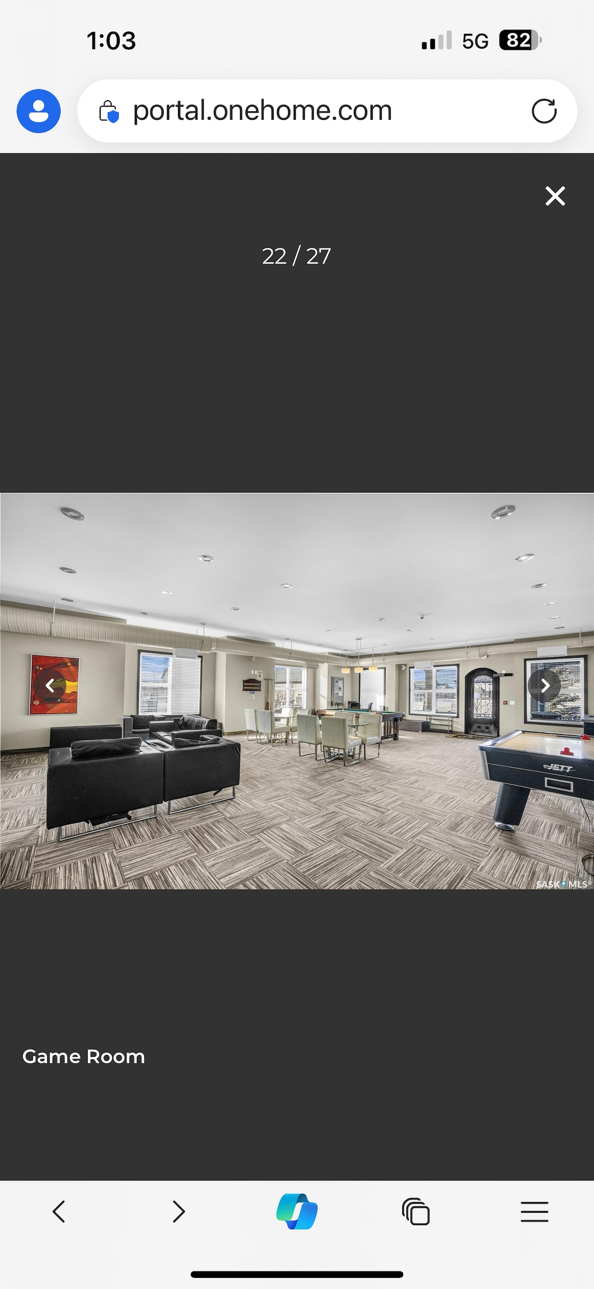 A spacious game room is seen with a variety of seating options arranged around modern furnishings. A pool table occupies one corner, while large windows allow natural light to flow in, illuminating the clean, neutral décor. A dining table is positioned nearby for gatherings and activities.