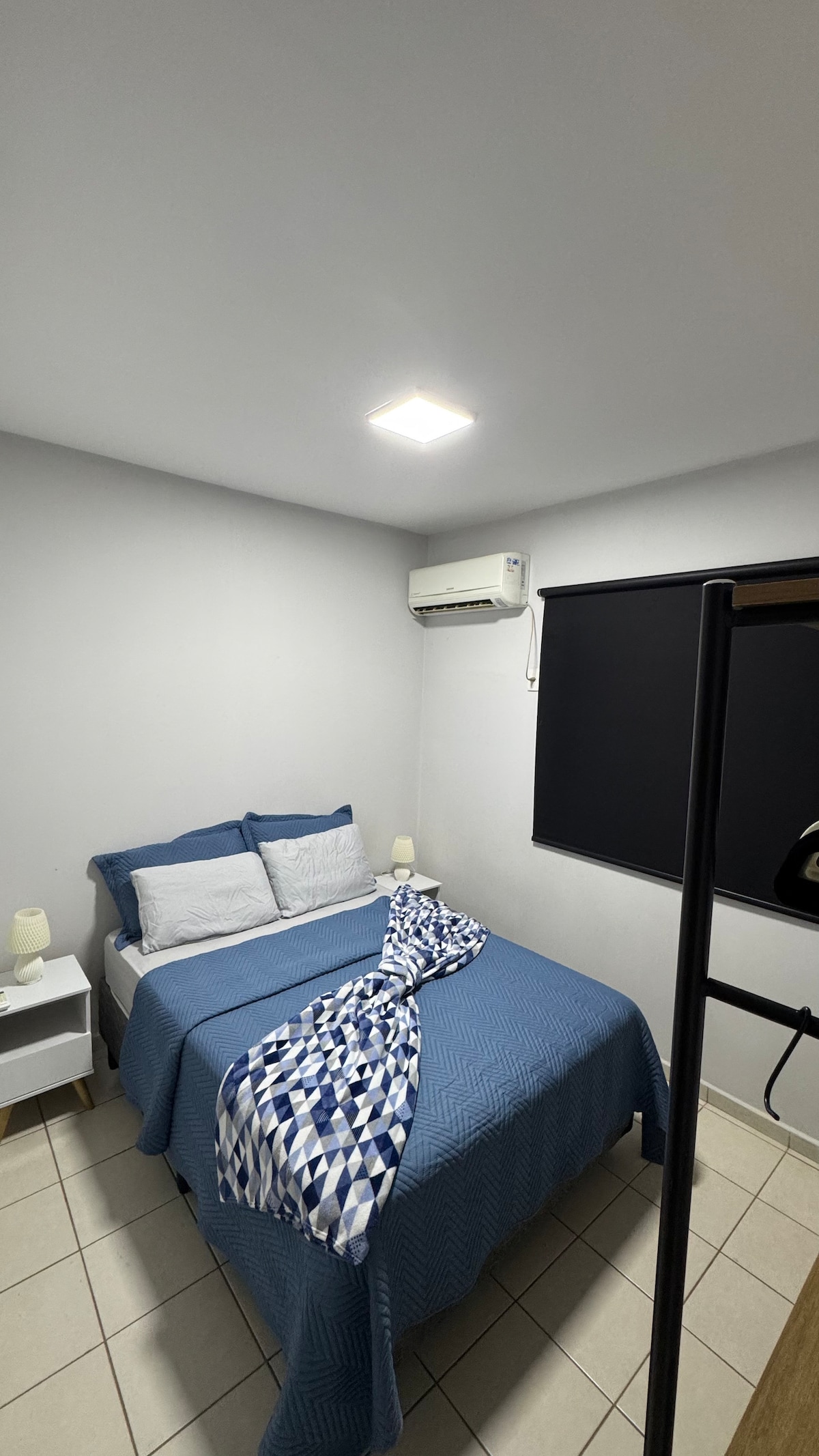 A cozy bedroom features a queen-sized bed dressed in blue and white bedding. Soft lighting is provided by a ceiling fixture, while a window with a dark blind allows for light control. A bedside table holds lamps for nighttime illumination.