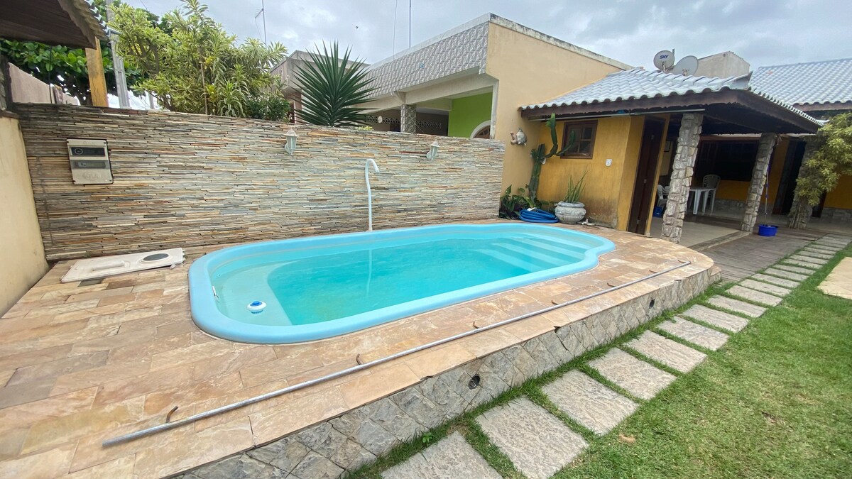 A tranquil outdoor space features a sparkling pool surrounded by a stone patio. Lush greenery adds to the ambiance, while a textured stone wall enhances the environment. A pathway of stepping stones leads to the grassy area, providing a seamless connection between spaces.
