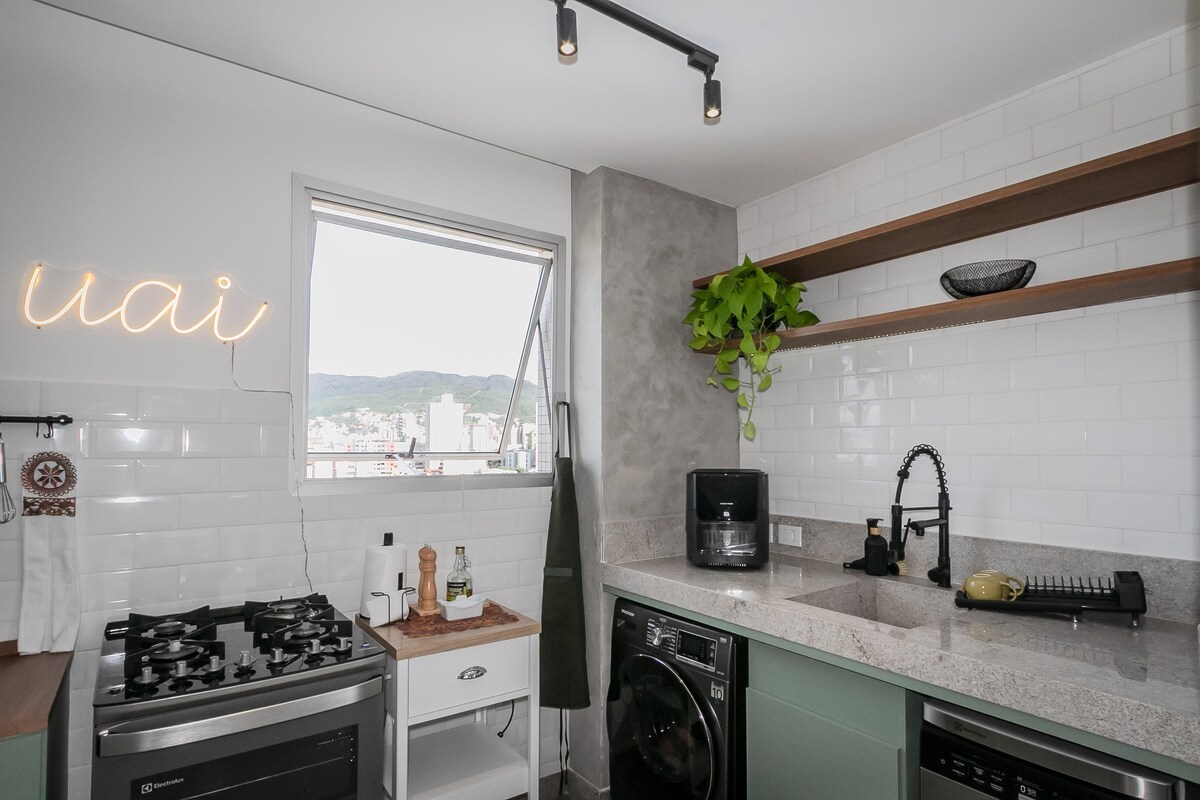 A modern kitchen area features light tile backsplash and a spacious counter with built-in appliances. Overhead shelves are adorned with decorative items and greenery. A large window offers a view of the surrounding mountains, allowing natural light to brighten the space.