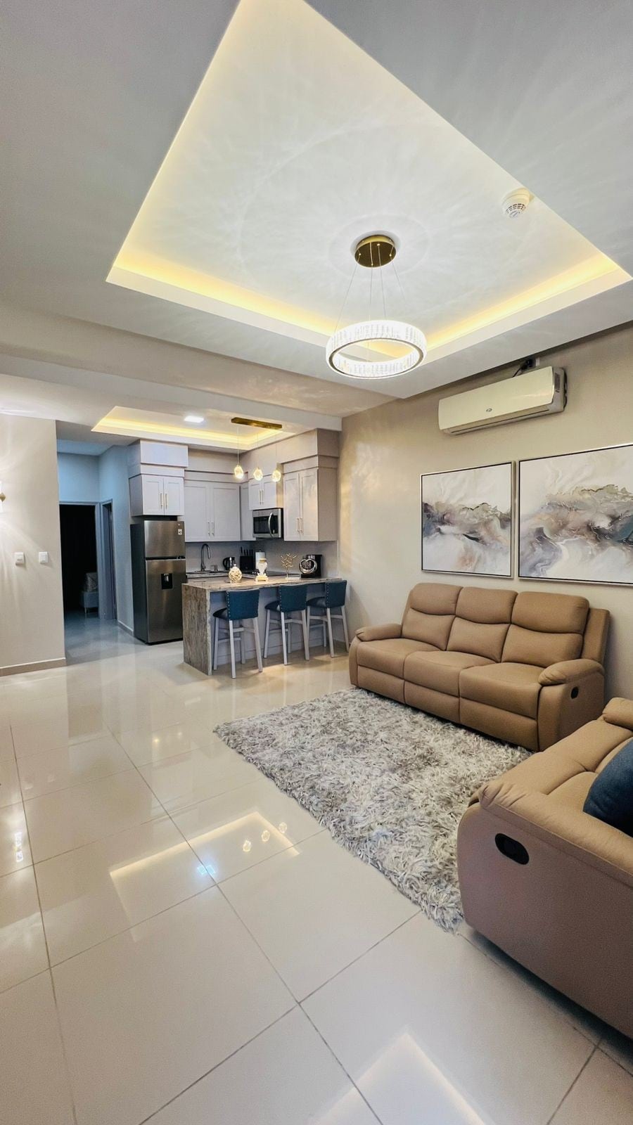 An open living space is presented with modern furnishings. A comfortable sofa and recliner are arranged on a plush area rug. The kitchen features sleek cabinetry and modern appliances. Soft lighting is provided by a unique ceiling fixture, enhancing the overall sophistication of the room.