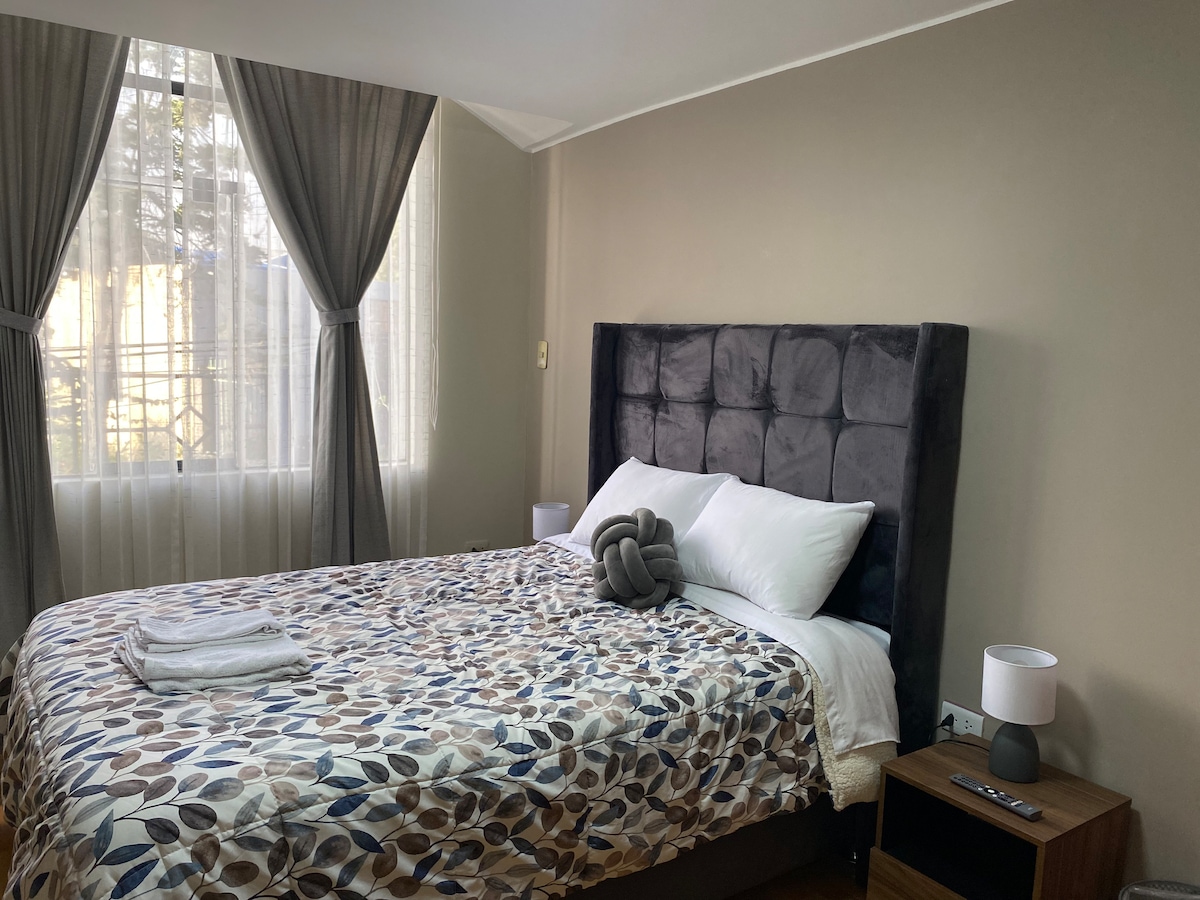 A spacious bedroom features a plush, upholstered headboard and a colorful patterned bedding set. Natural light filters through sheer curtains, illuminating the room. A bedside table with a lamp and neatly arranged towels adds to the room's functionality.