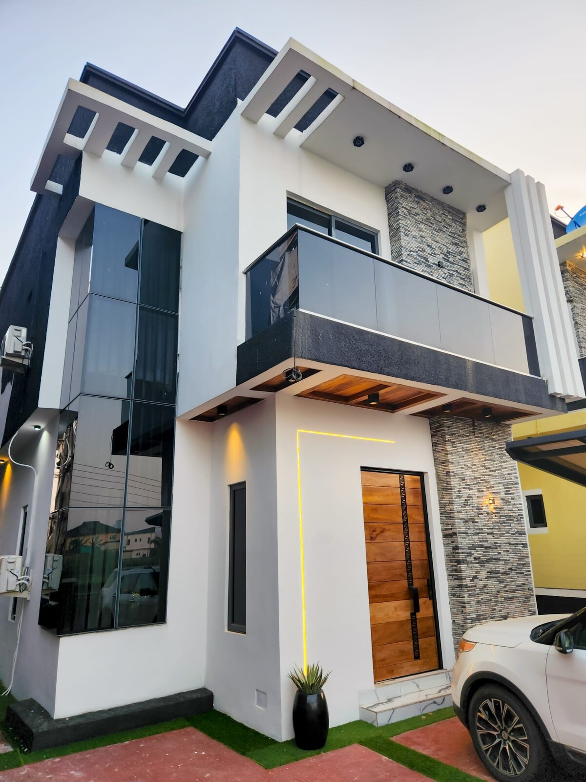 A modern two-story home features a striking facade combining sleek glass and textured stone. A wooden door, framed by a simple planter, adds a touch of warmth. The balcony showcases clean lines and a minimalist design, enhancing the overall contemporary aesthetic.