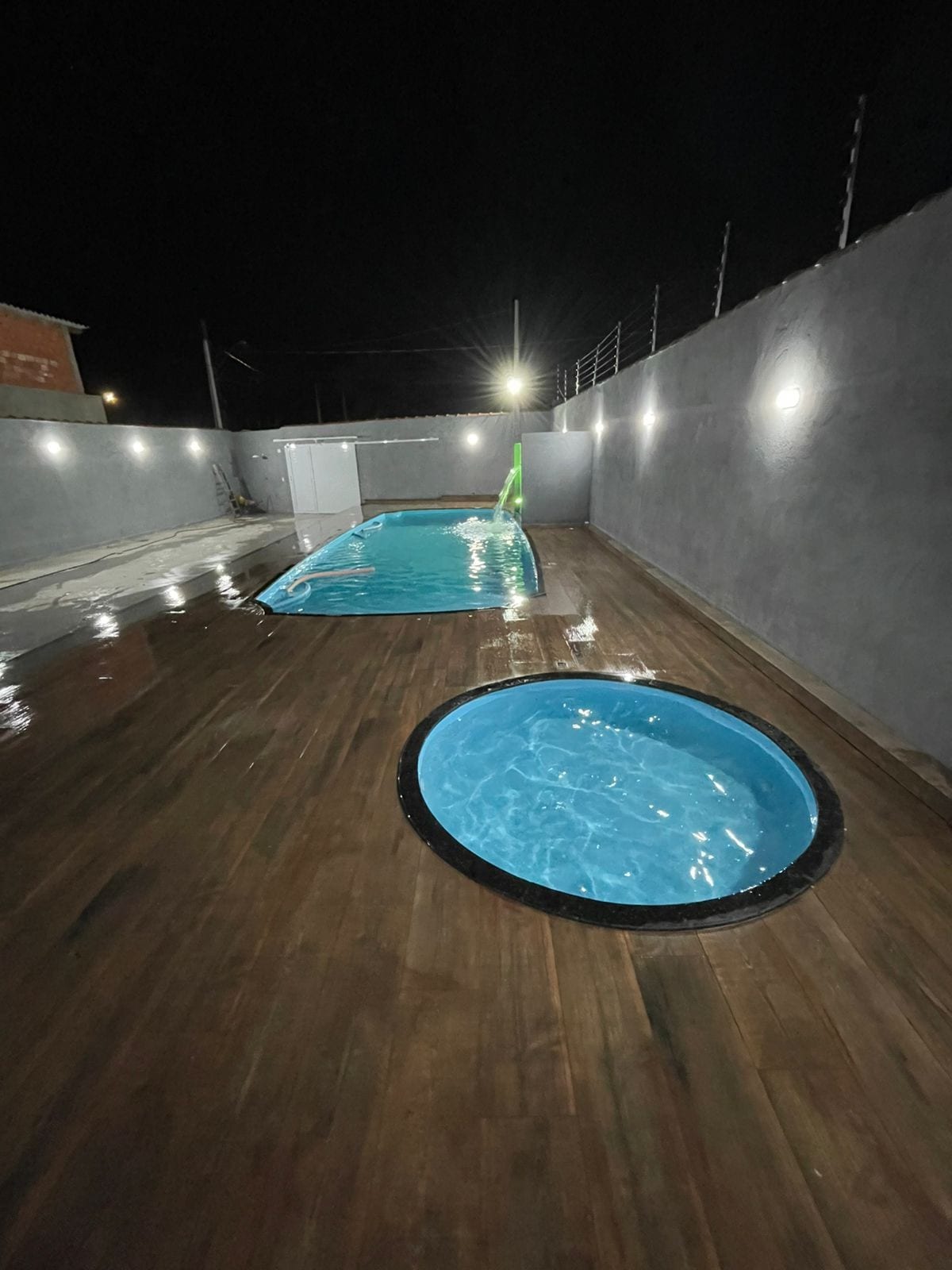A well-lit outdoor area features two pools at night, one rectangular and larger, and a smaller round spa. The surrounding wood deck reflects the light, enhancing the inviting atmosphere. Walls are painted in a light tone, and soft lighting highlights the space.