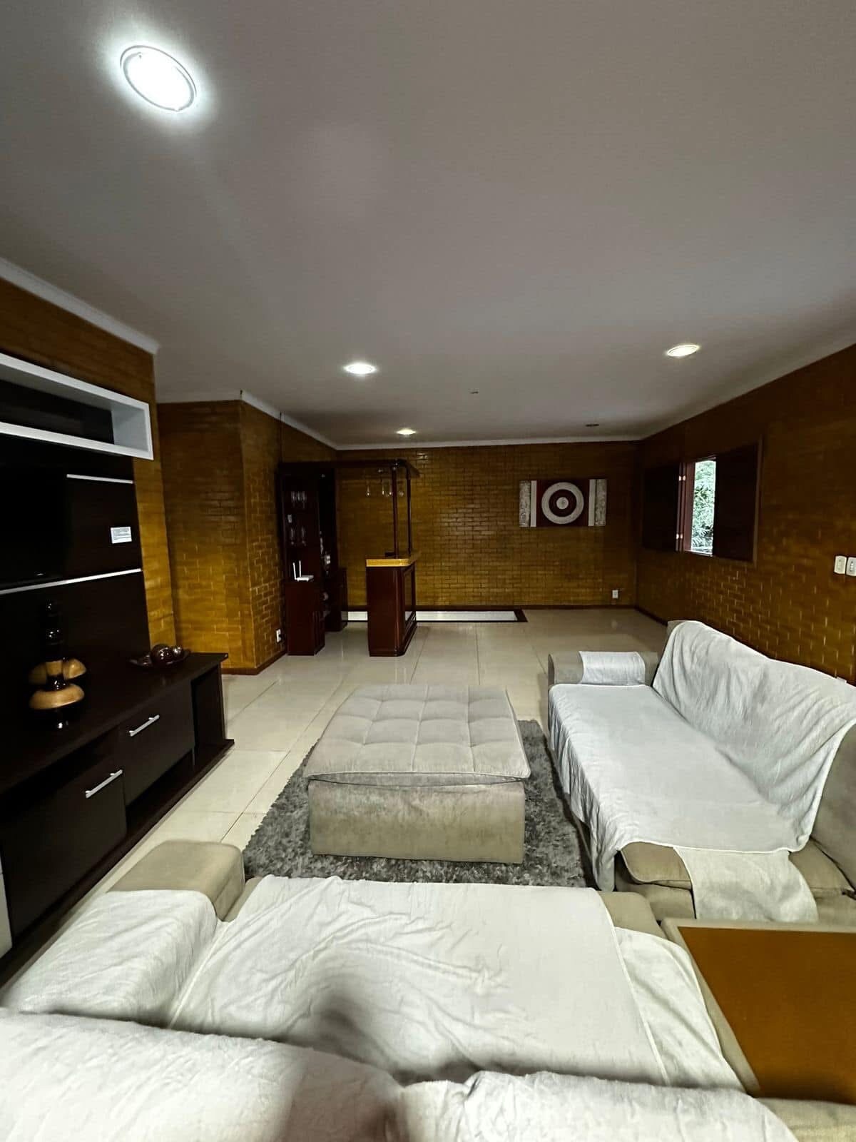 The living area features light-colored sofas arranged around a central ottoman, with warm walls creating a cozy atmosphere. A wall unit displays decorative items, while a doorway leads to an adjoining space. Natural light enters through nearby windows, enhancing the room's openness.