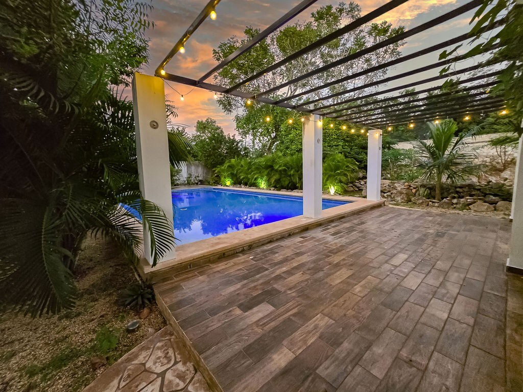 An outdoor space features a tranquil pool surrounded by lush greenery. Soft ambient lighting is provided by string lights hanging above. A wooden deck area leads to the pool, which reflects the warm hues of a sunset sky.