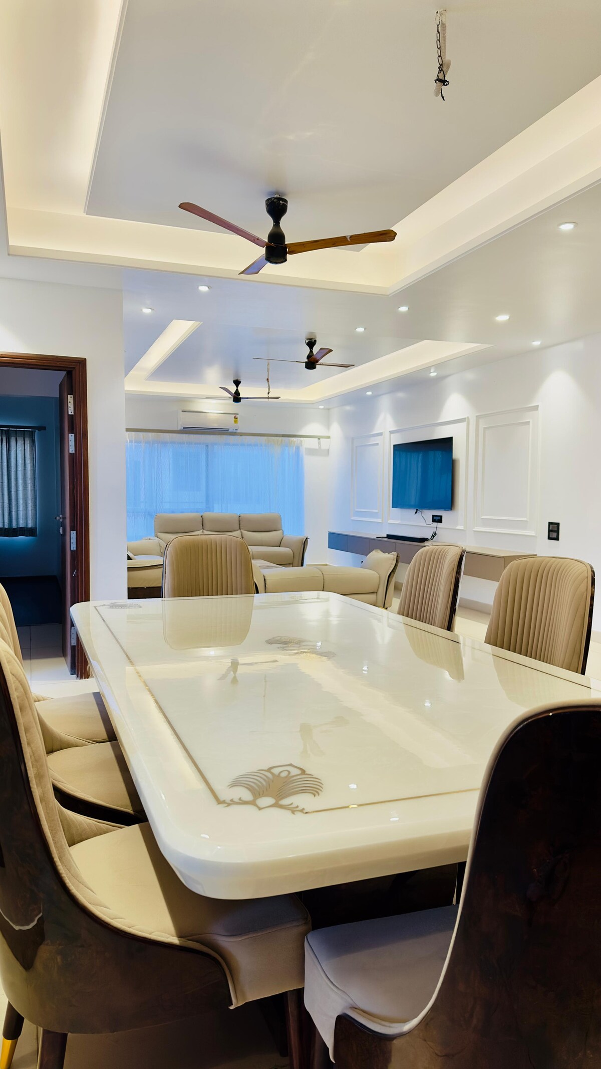 A modern dining area features a large glass-top table surrounded by beige and dark wood chairs. Ceiling lights and fans enhance the bright ambiance, while a sleek television is mounted on the wall, offering an inviting space for gatherings.