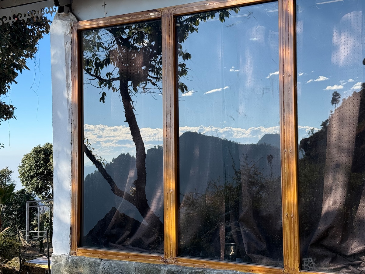 Large windows feature wooden frames, offering a view of the mountains and valley beyond. Reflections of trees and clouds are visible in the glass, while sheer curtains partially cover the panes, allowing filtered light to enter the space.