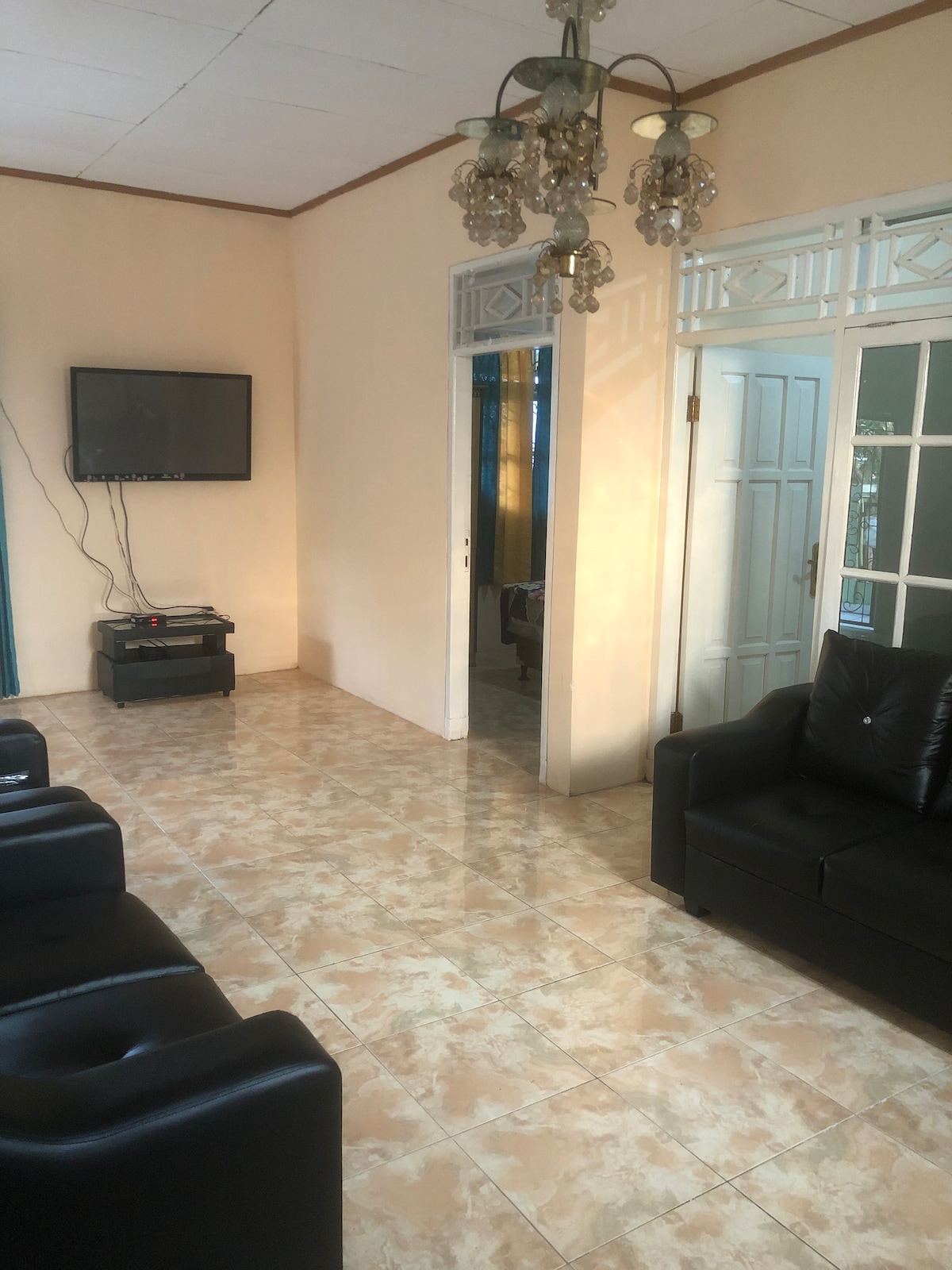 A living area features tiled flooring and walls painted in soft tones. Two black leather sofas provide seating, facing a mounted television. A glass door leads to another space, illuminated by natural light coming from nearby windows.
