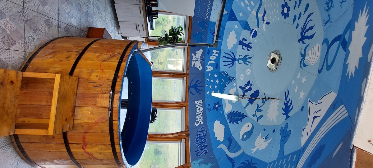 A large wooden hot tub is prominently featured, surrounded by a blue mural depicting oceanic scenes. Natural light streams through the tall windows, illuminating the space and enhancing the blue tones of the artwork and tub.