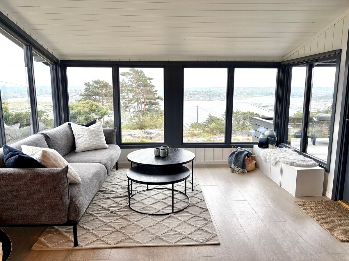 A sunlit living area features large windows offering panoramic views of the surrounding landscape. A cozy grey sofa is accompanied by a circular black coffee table. A patterned area rug adds texture to the wooden floor, while decorative elements are positioned thoughtfully around the space.