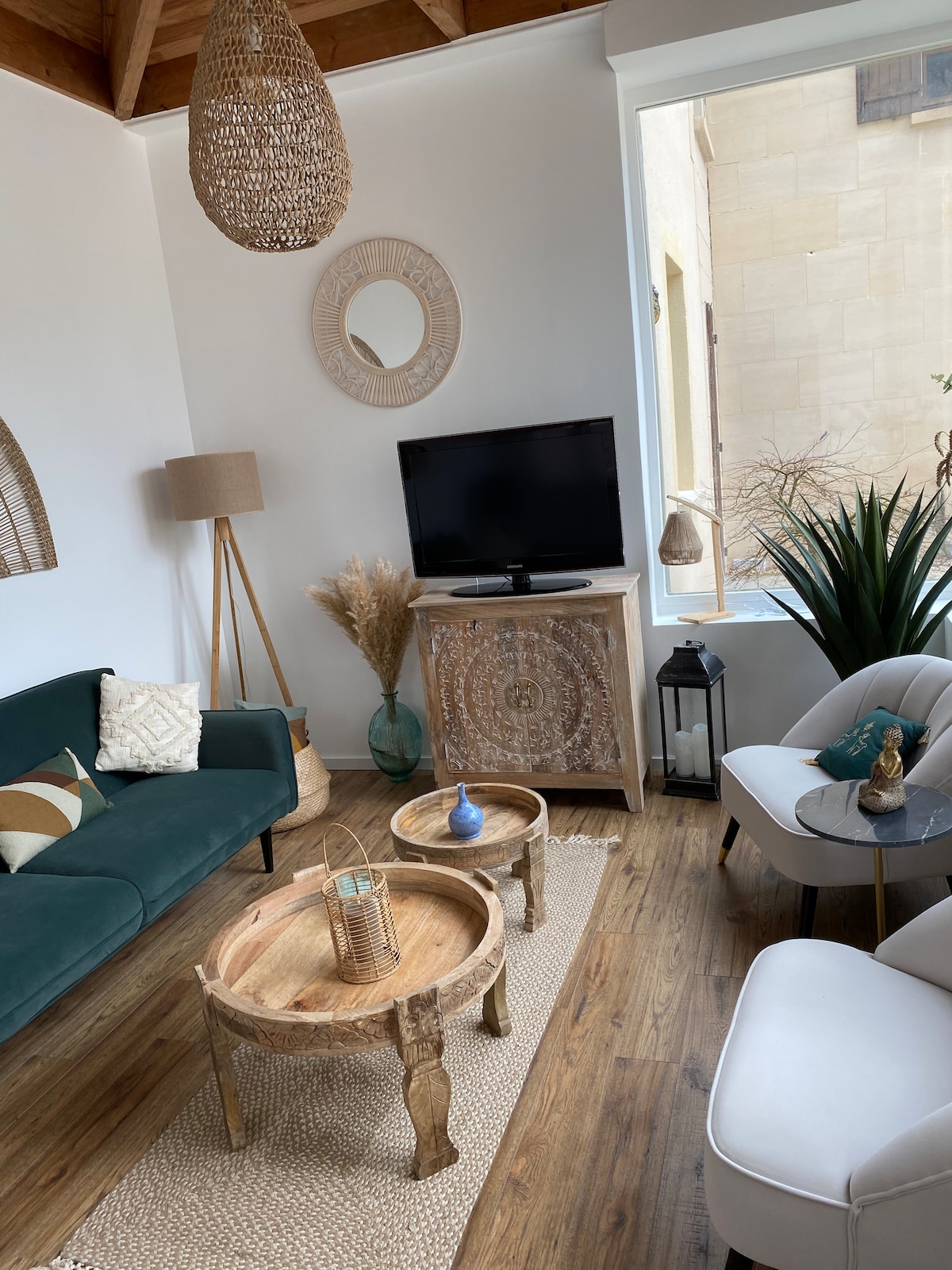 A cozy living area features a green sofa with decorative pillows and a small side table. Two round wooden tables sit on a natural fiber rug. A TV is mounted above a decorative cabinet, while various plants and light fixtures add warmth to the space.