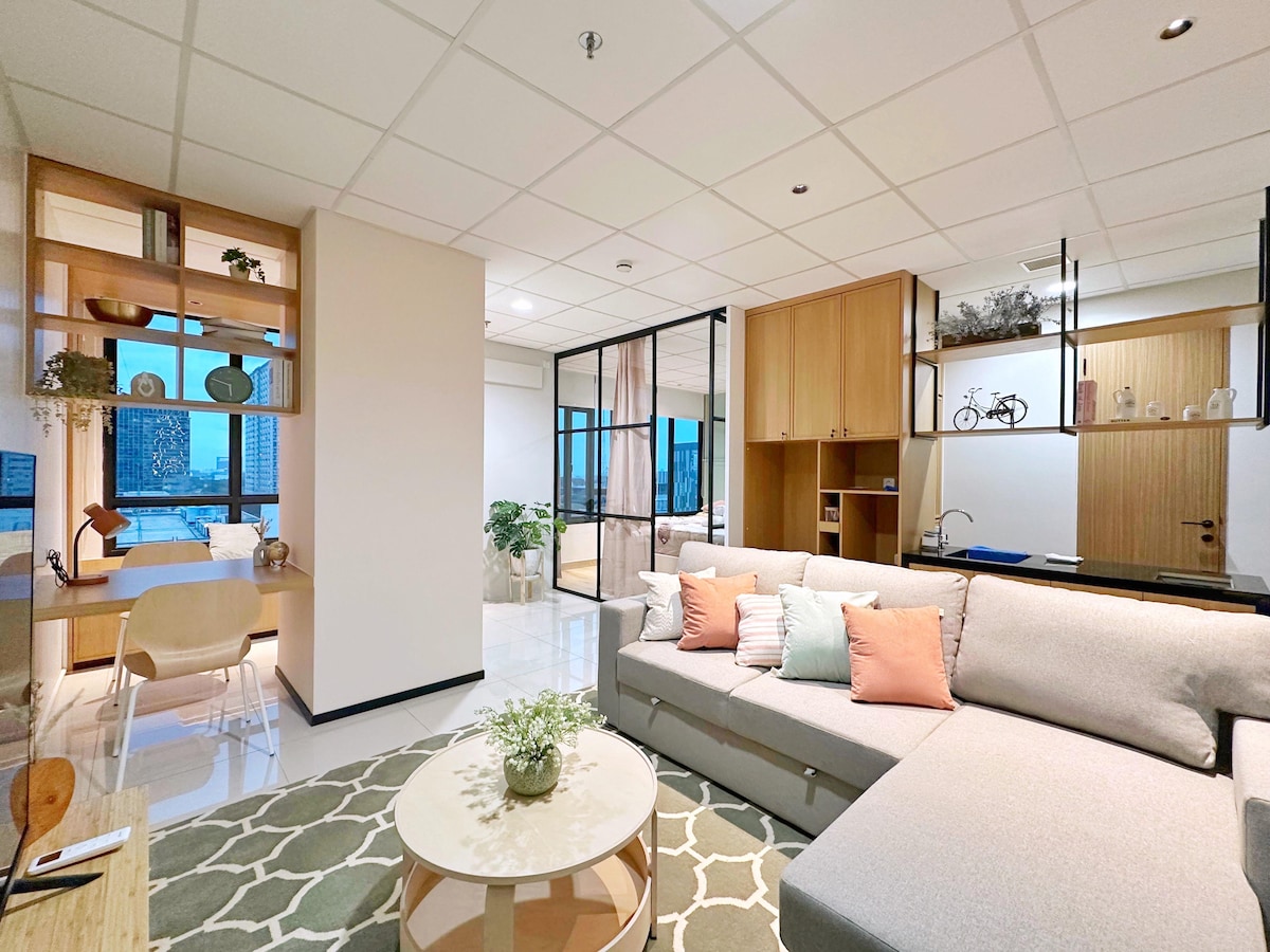 The living area features a spacious light-colored sofa adorned with decorative pillows, complemented by a round coffee table with a small potted plant. A modern work station with a desk and chair is visible, alongside large windows providing views of the cityscape.