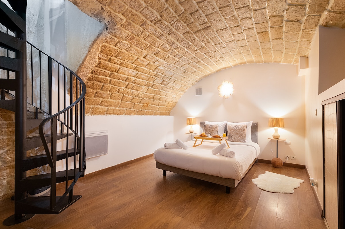 A spacious bedroom is showcased, featuring a contemporary design with an exposed stone arch ceiling. A comfortable bed is centered within the room, accompanied by two bedside tables and lamps. A spiral staircase leads to an upper level, enhancing the unique layout of the space.