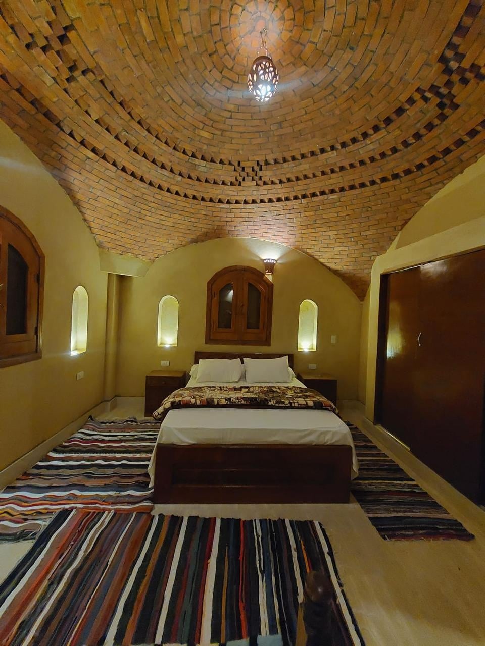 A cozy sleeping area features a spacious bed with decorative bedding. The room is enhanced by a unique brick ceiling and warm lighting. Traditional rugs are placed on the floor, complementing wooden furnishings and soft wall colors.
