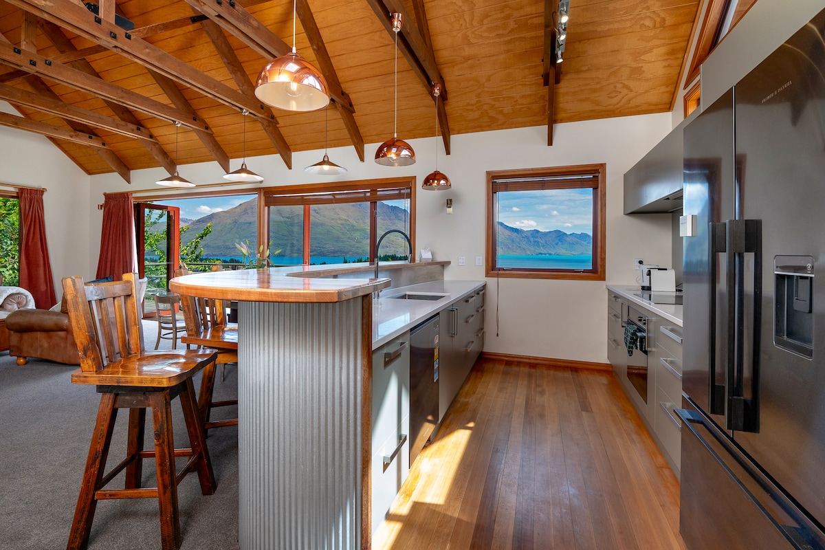 A modern kitchen features sleek cabinetry and a large central island with bar seating. Natural light enters through large windows that frame stunning views of the mountains and lake. Wooden beams and a warm color palette contribute to the inviting atmosphere.