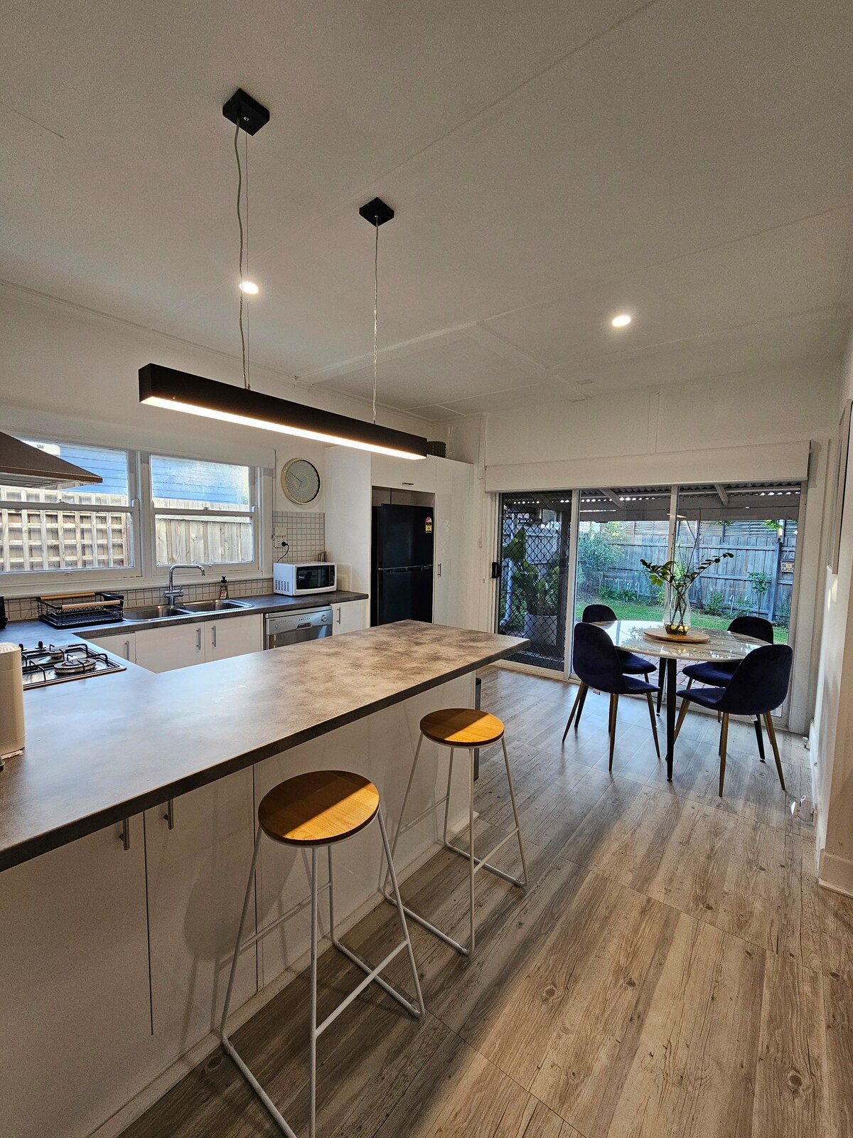 A modern kitchen features a central island with two high stools and a sleek countertop. Large sliding doors open to a cozy outdoor space filled with greenery. Natural light brightens the area, which includes a round dining table set for four, enhancing the inviting ambiance.