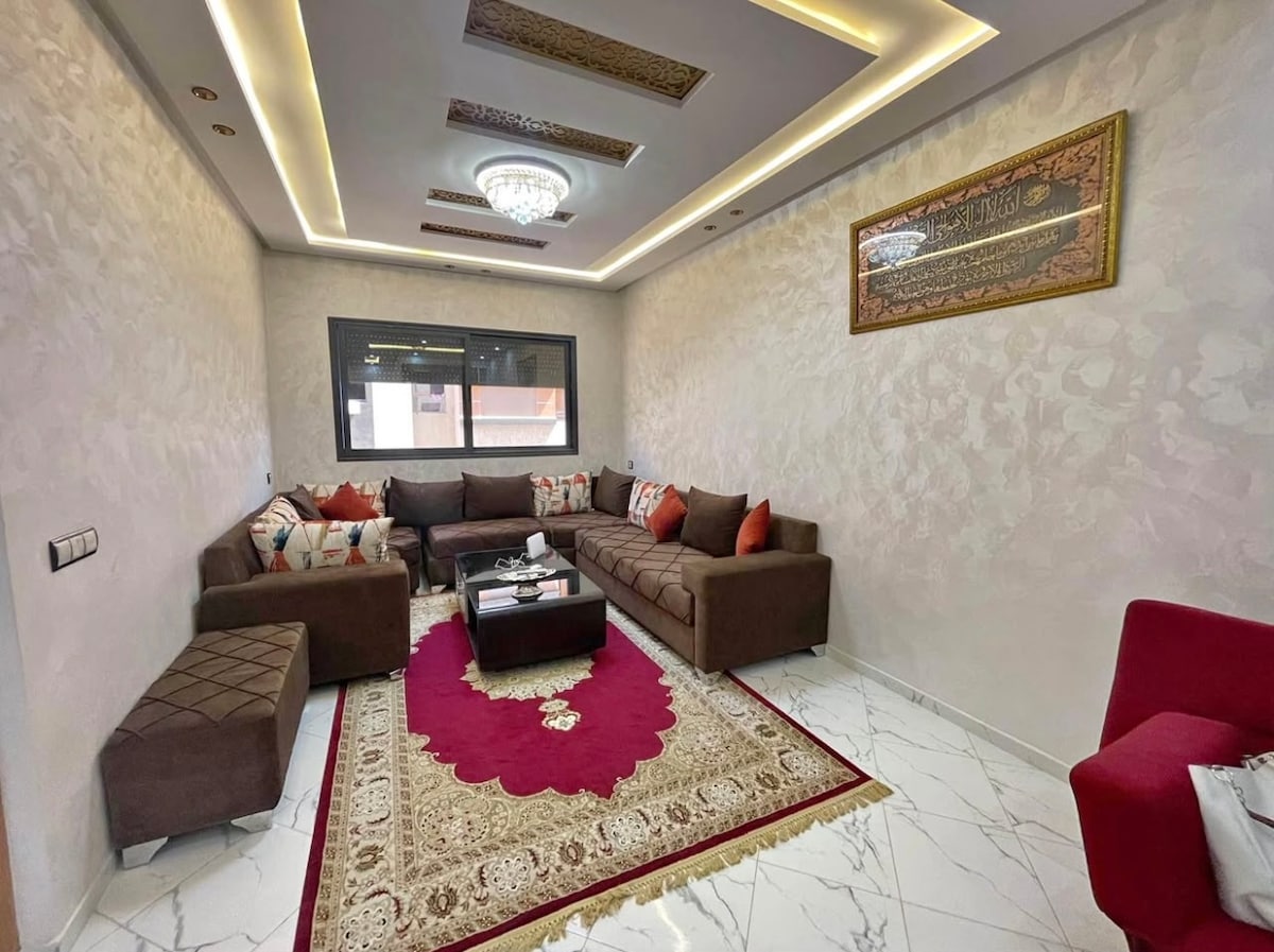 A comfortable living area features a large brown sectional sofa adorned with colorful cushions. A dark wood coffee table sits at the center, complemented by a decorative rug with intricate patterns. Soft lighting enhances the clean design, with windows providing natural light.