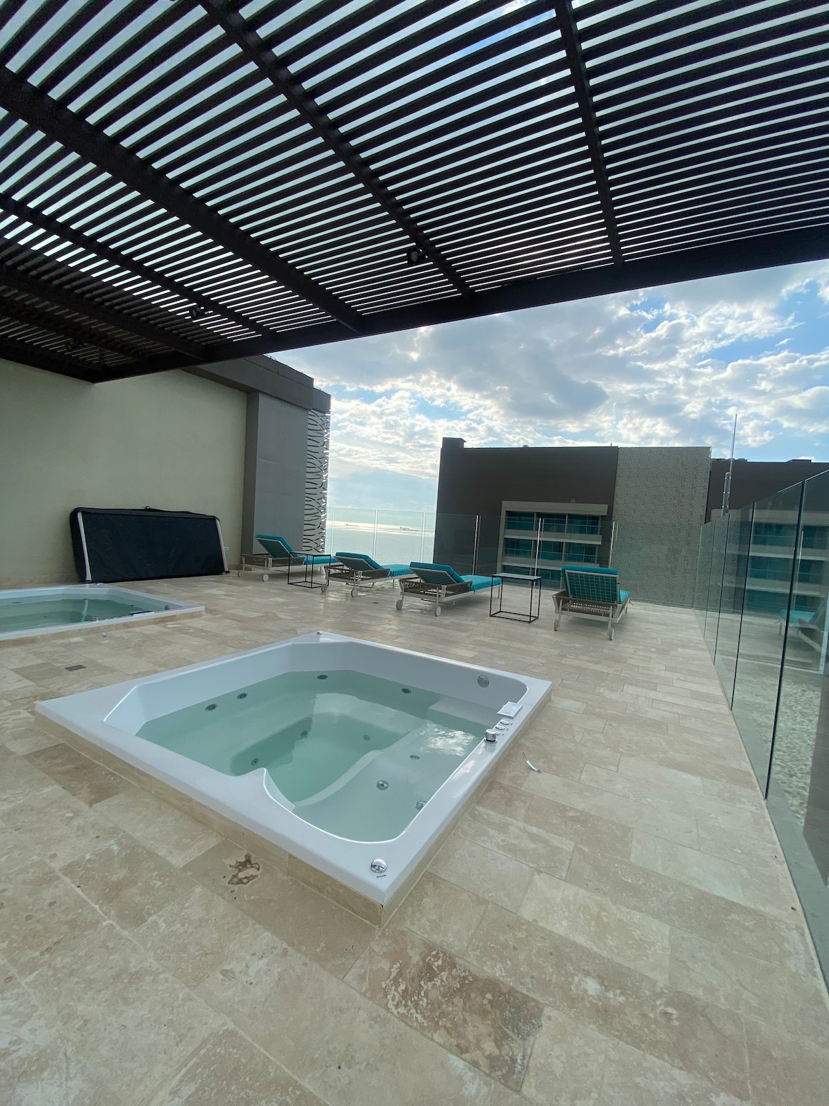 An outdoor terrace features a jetted tub surrounded by neutral stone tiles. Comfortable teal lounge chairs are positioned nearby, with a partial view of the ocean in the background. The area is covered by a slatted pergola, offering a shaded space to relax.