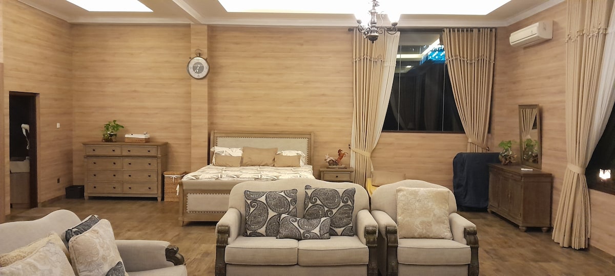 A spacious bedroom features warm wood paneling and soft lighting. A large bed is positioned beside a window dressed with flowing curtains. Comfortable seating is arranged around a coffee table, with a dresser and mirror nearby, creating a welcoming atmosphere for relaxation.