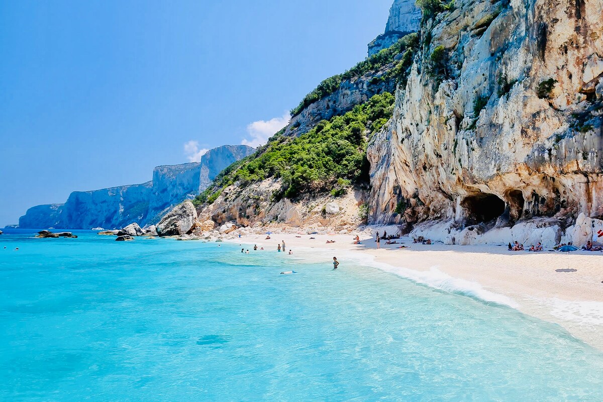 A serene beach scene is depicted, featuring soft white sand and clear turquoise waters. Gentle waves lap at the shore, while rocky cliffs provide a natural backdrop. Sunbathers and visitors are scattered along the beach, enjoying the warm, sunny day.