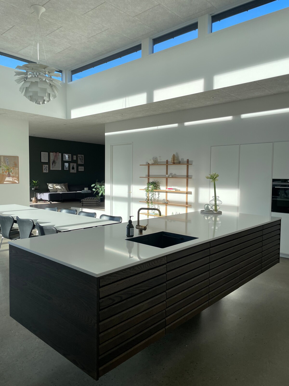 A modern kitchen island with a dark wood finish is centered in the space, featuring a sleek white countertop. Natural light filters through large windows, illuminating a minimalist design with potted plants and wooden shelving in the backdrop. An open layout connects to a living area.