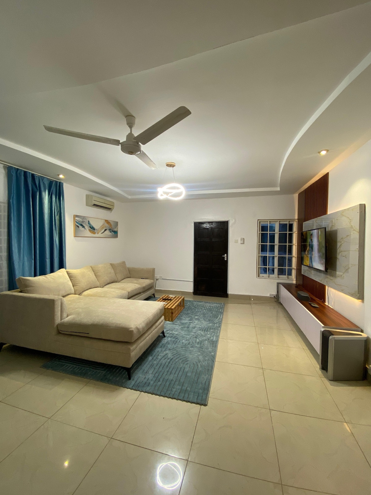 A comfortable living area features a light-colored sectional sofa arranged around a low wooden coffee table. A plush area rug sits on the polished tile floor. A wall-mounted television is positioned against a light-colored feature wall, while a ceiling fan and elegant lighting provide a well-lit ambiance.