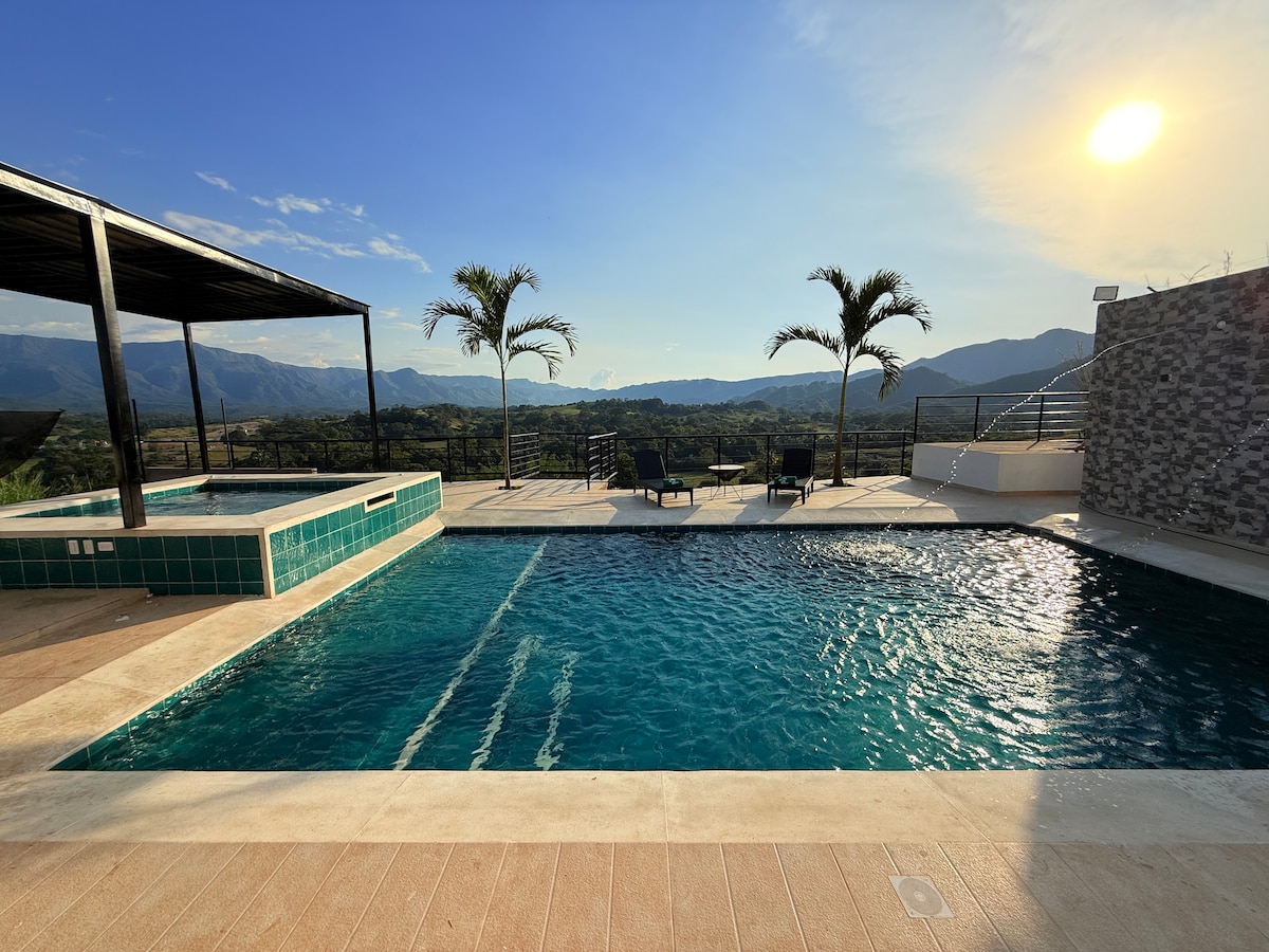 A spacious pool area is featured, including a private swimming pool and a jacuzzi, surrounded by natural landscapes. Palm trees gently sway in the background, and the sun sets beyond the distant mountains, casting a warm glow over the tranquil scene.
