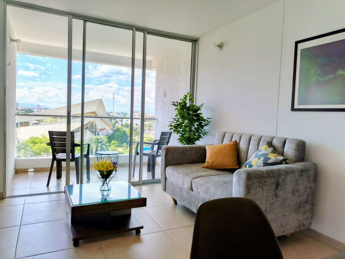 A bright living area features a gray sofa adorned with decorative pillows, positioned next to a glass coffee table. A potted plant adds a touch of greenery. Floor-to-ceiling glass doors open to a balcony with outdoor seating, offering views of the surrounding landscape.