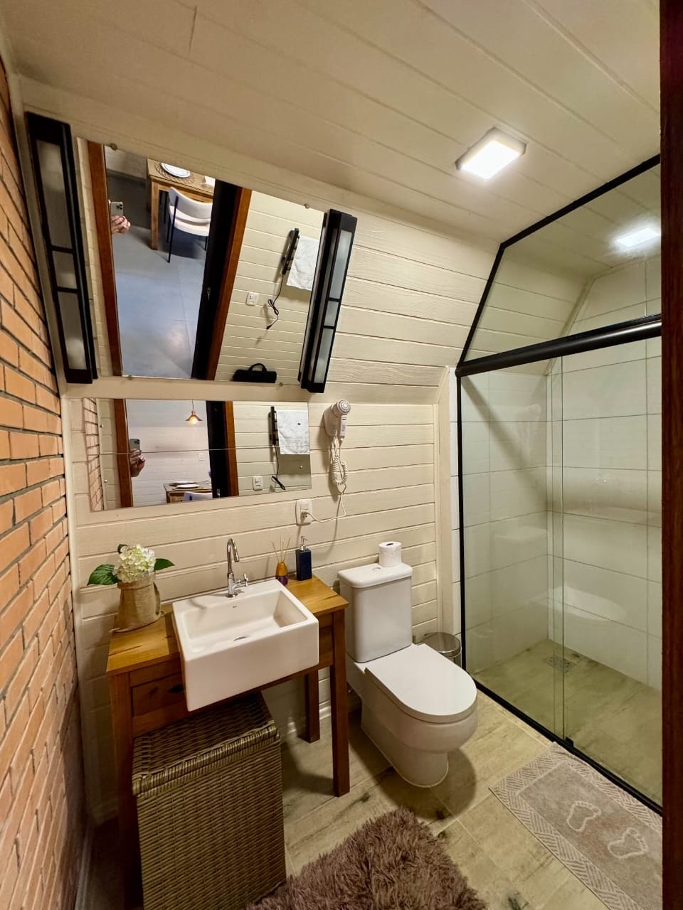 The bathroom features a modern sink and a window providing natural light. A glass shower enclosure is visible, alongside a toilet and a small woven storage basket. Two mirrors enhance the sense of space, while warm wooden accents add a cozy touch.