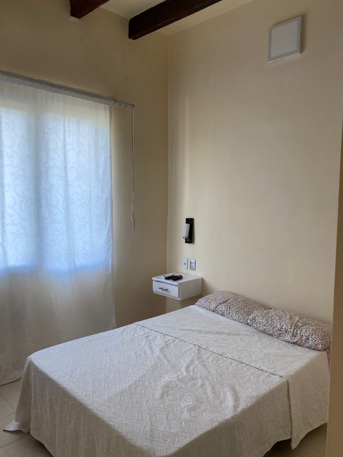 A simple sleeping area features a spacious bed covered with a white bedspread. A small nightstand is positioned beside the bed, equipped with a telephone and a drawer. Natural light filters through a sheer curtain covering the window.
