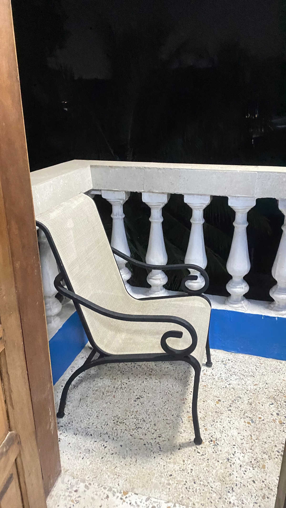 An outdoor balcony area is featured, showcasing a wrought iron chair positioned against a traditional balustrade. The concrete flooring is visible, with a low wall providing a partial view of the surrounding greenery under night lighting.