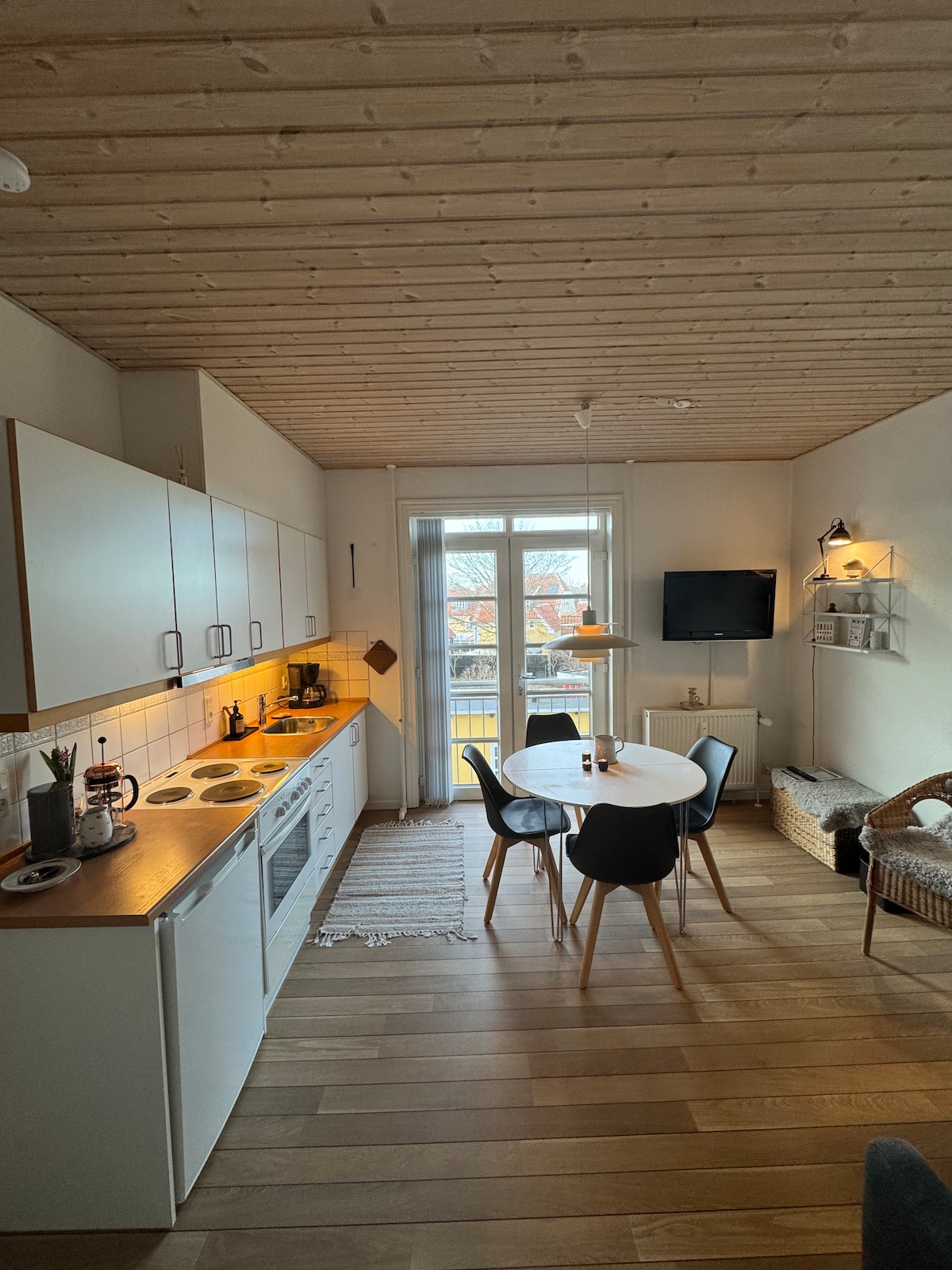 An inviting kitchen and living area is showcased, featuring light wood flooring and a neutral color palette. A dining table with four chairs is positioned near a window, and a compact kitchen includes essential appliances. A television is mounted on the wall, with natural light enhancing the space.