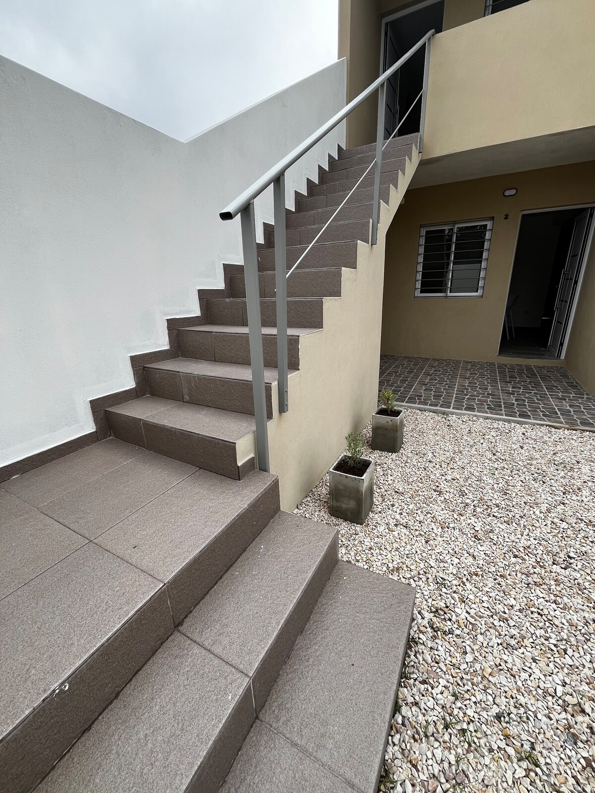 An exterior view showcases a set of stairs leading upward, flanked by light-colored walls and a modern metal railing. Small planters with greenery are positioned at the base of the stairs, while a paved area is visible to the side.