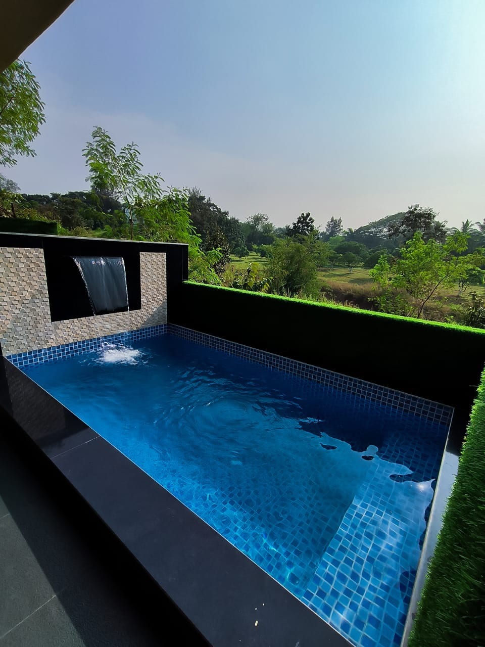 A tranquil swimming pool is depicted, featuring clear blue water reflecting the surrounding greenery. A gentle waterfall feature provides a soothing ambiance. Lush trees and open fields are visible in the background, enhancing the serene outdoor setting.