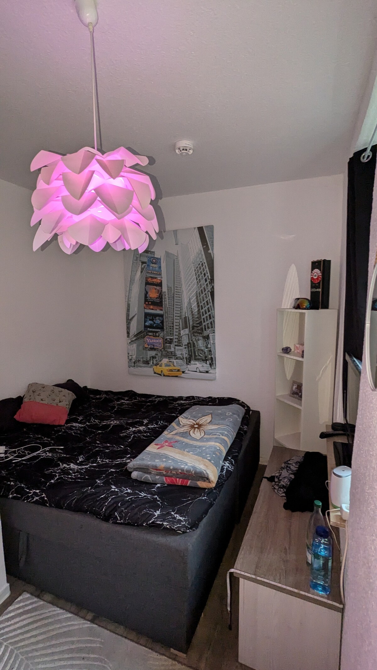 A cozy sleeping area features a spacious bed adorned with a striped blanket and decorative pillows. A wall-mounted poster adds character, while a unique light fixture casts a soft glow. A wooden shelf displays various items, contributing to the room's functional design.