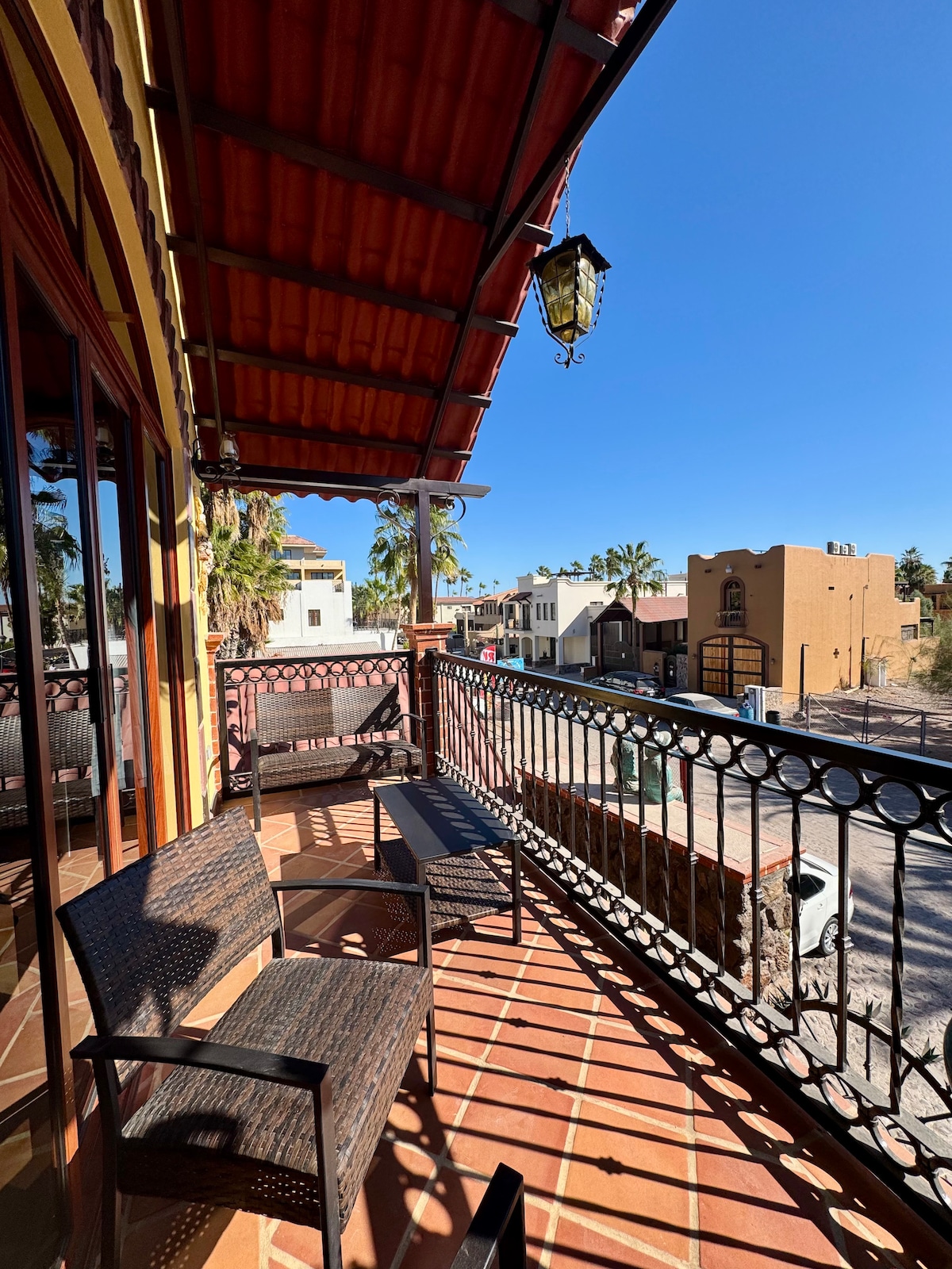 A balcony area is shown with two wicker chairs and a small table. The tiled flooring and wrought iron railing complement the surrounding architecture. Bright blue skies are visible above, with a glimpse of neighboring buildings and palm trees in the background.