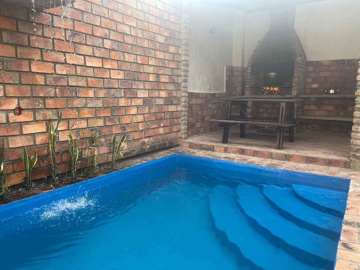 A vibrant blue pool with gentle steps leads into the water, surrounded by rustic brick walls. A small garden area features green plants along the edge. In the background, a traditional brick barbecue area is visible, complemented by a wooden table and benches.