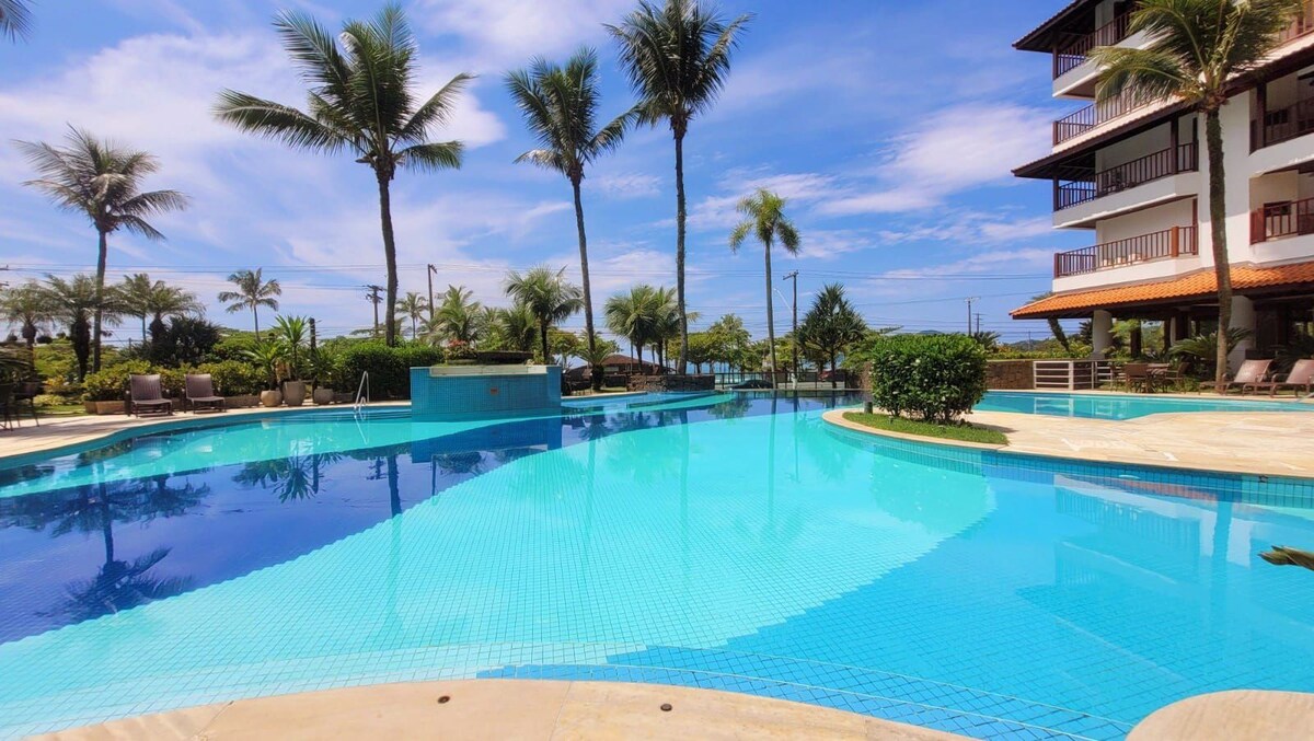 A serene pool area is surrounded by lush palm trees and vibrant greenery. The clear blue water reflects the sky, while comfortable lounge chairs are positioned along the water's edge, inviting relaxation. A gentle breeze can be seen through the surrounding landscape.