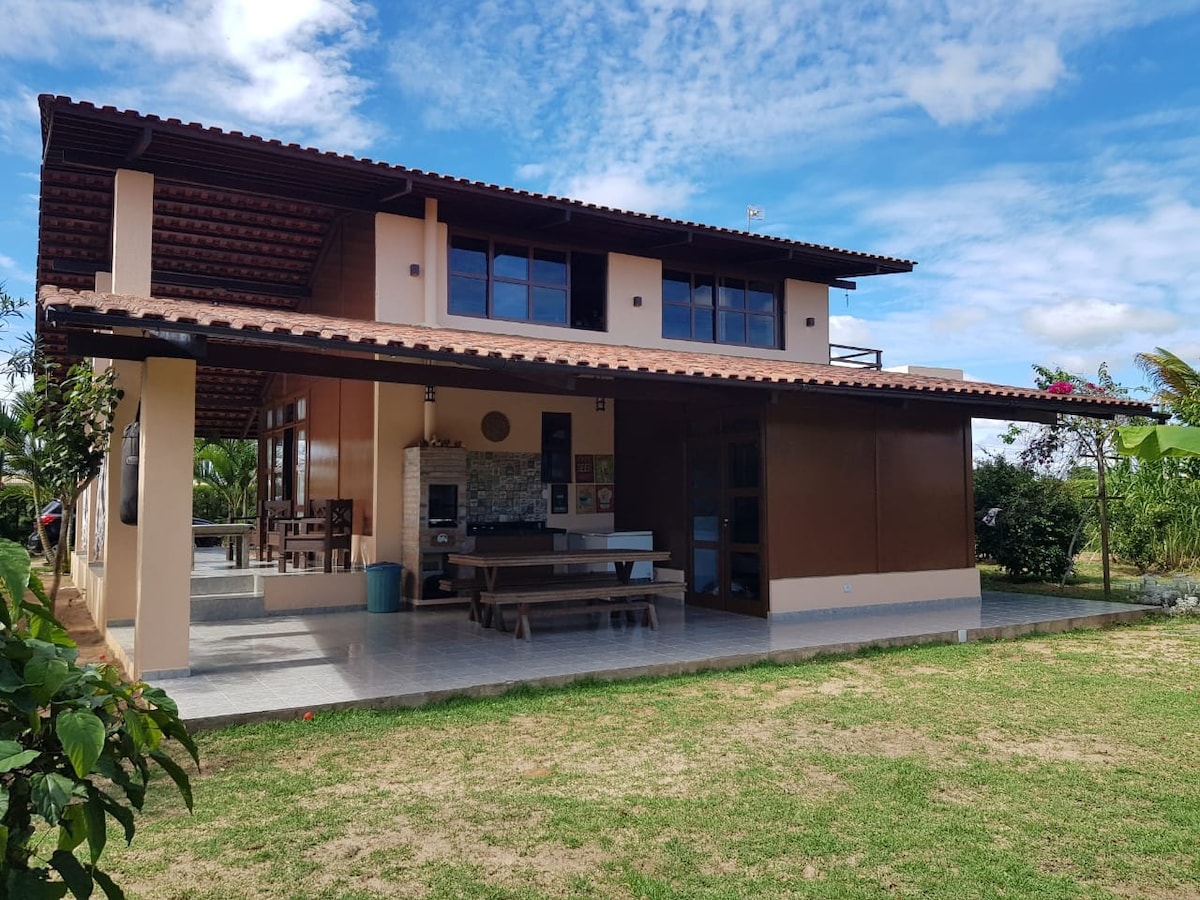 The exterior of a two-story house is shown, featuring a spacious covered terrace with a large dining table. Large windows allow natural light into the interior. A well-maintained lawn and garden area are present, along with trees and greenery in the background.